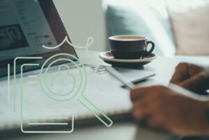 Ein laptopt auf einem Tisch, auf dem eine Kaffeetasse steht und zwei Hände vor dem Laptop auf ein Blatt schreiben.