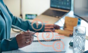 Eine Person am Schreibtisch, von der man nur die Hände sieht, wie sie etwas schreiben zum Thema Recruiting 2025 Warum Priorisierung im Fachbereich der unterschätzte Schlüssel ist. Außerdem ist ein Laptop auf deiner Docking Station zu sehen und ein Glas Wasser