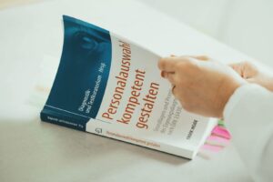 ein buch über eignungsdiagnostik liegt auf einem Tisch und wird von zwei Händen durchgeblättert. Es beschäftigt sich mit der Frage: Wie ticken Bewerber:innen wirklich?